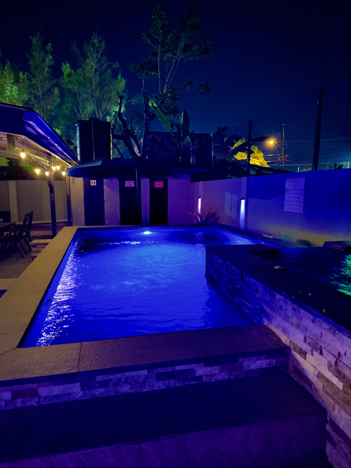 A private backyard pool is showcased, illuminated in calming blue tones under the night sky. Surrounding the pool, there is a stone wall and soft lighting that creates a serene ambiance. An outdoor seating area is visible nearby, enhancing the outdoor space.
