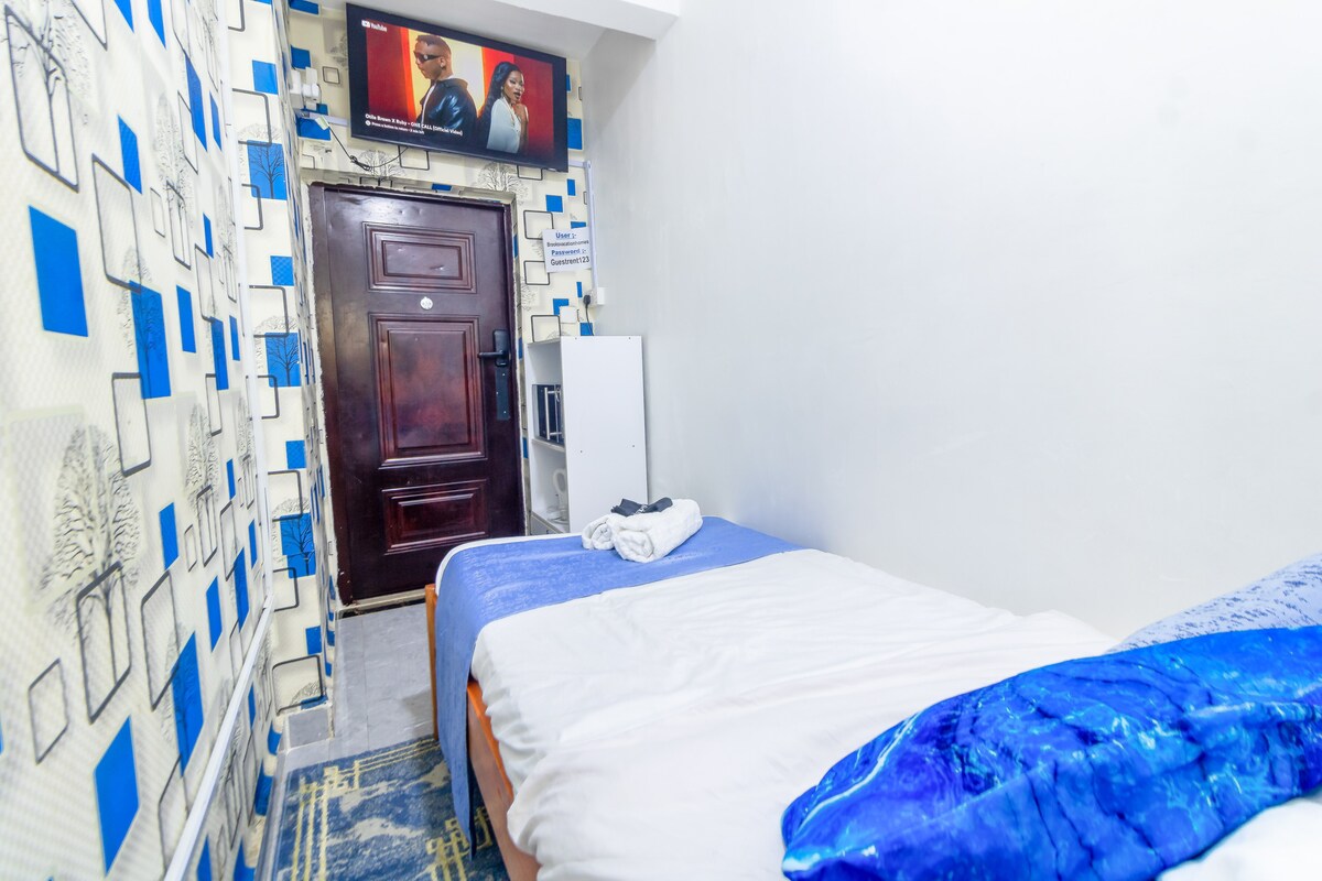 The room features a snug sleeping area with a spacious bed covered in white linens and a blue throw. A 50-inch smart TV is mounted on the wall, and a private door leads to the outside. The walls are adorned with a modern blue and white design.