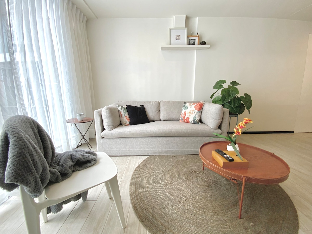 A light-filled living area features a spacious sofa adorned with colorful cushions. A round coffee table sits at the center, complemented by a decorative plant nearby. Sheer curtains allow natural light to filter in, creating a warm ambiance throughout the space.