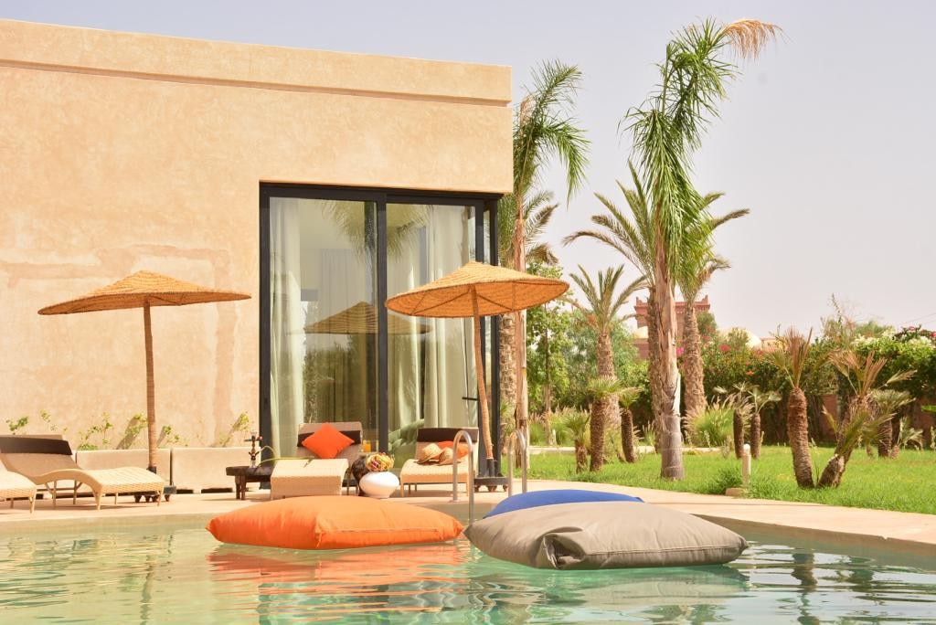 A serene outdoor space features a pool with colorful floating loungers. Palm trees provide shade from large umbrellas, while sunbeds sit nearby, offering a relaxing environment. Expansive glass doors reflect the lush garden beyond, connecting the outdoor area to the interior.