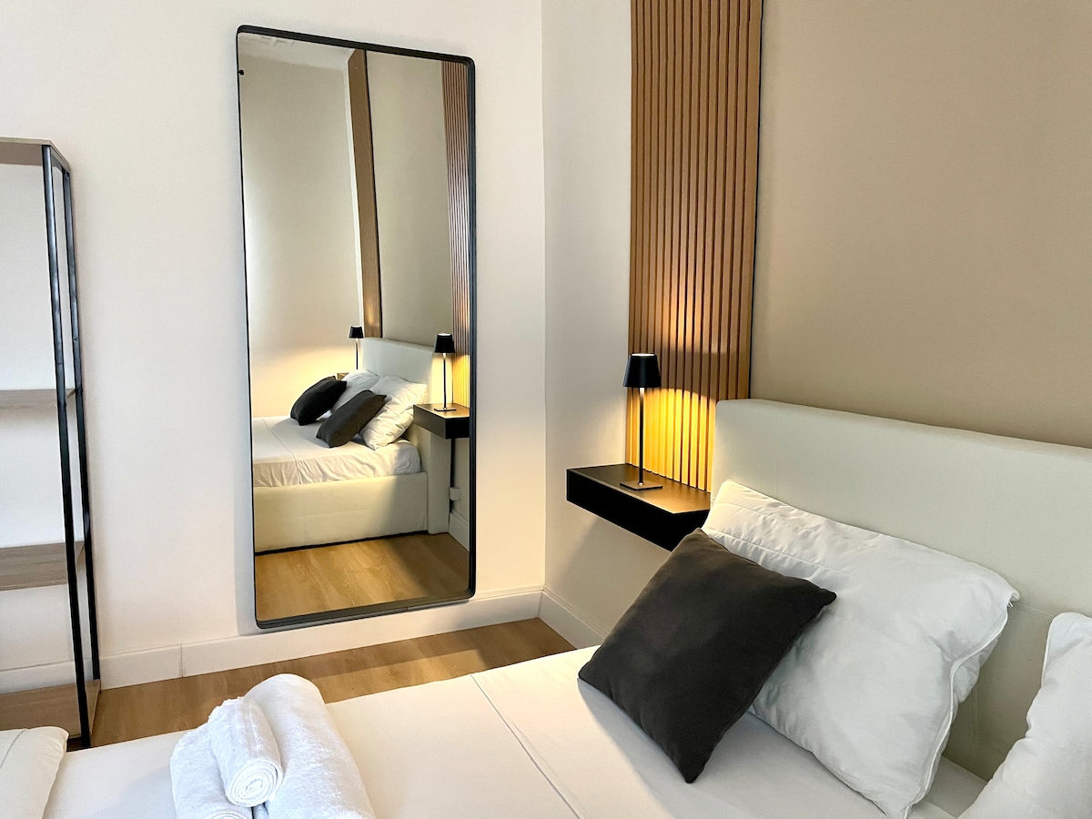 A spacious bedroom features a large mirror reflecting the comfortable bed, adorned with soft pillows and a cozy blanket. Elegant bedside lamps provide soft lighting, while a minimalist shelf showcases simplicity and functionality. Natural light enhances the warm tones of the decor.
