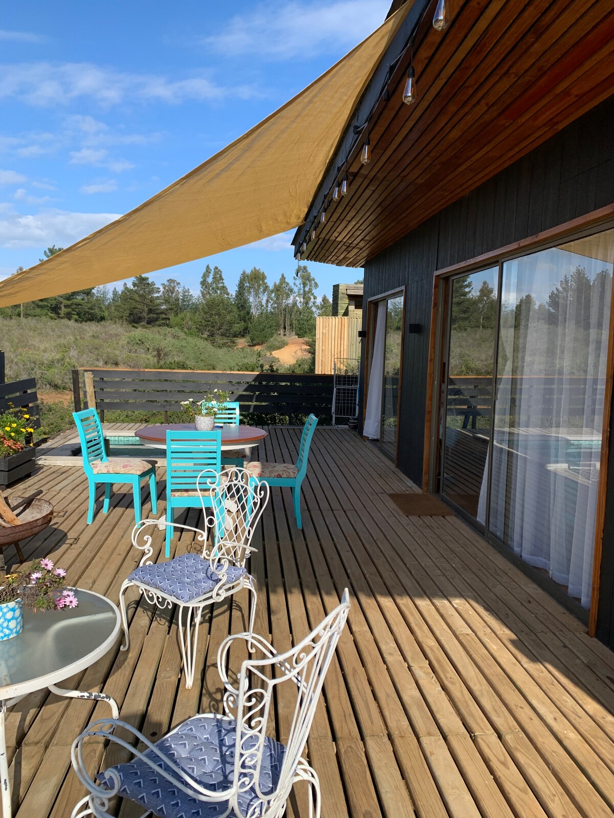 A spacious outdoor deck features a combination of table and chair sets in vibrant colors, complemented by elegant metal chairs. Sunlight filters through a large awning, creating shaded areas for relaxation. Potted flowers add color to the setting, surrounded by natural greenery.