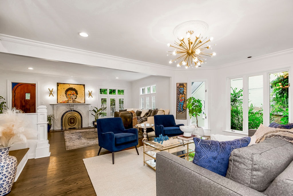 The open-concept living area features a mix of comfortable seating, including navy chairs and a grey sofa. A stylish chandelier provides overhead lighting. Large windows allow natural light to fill the space, with a view of lush greenery visible outside.