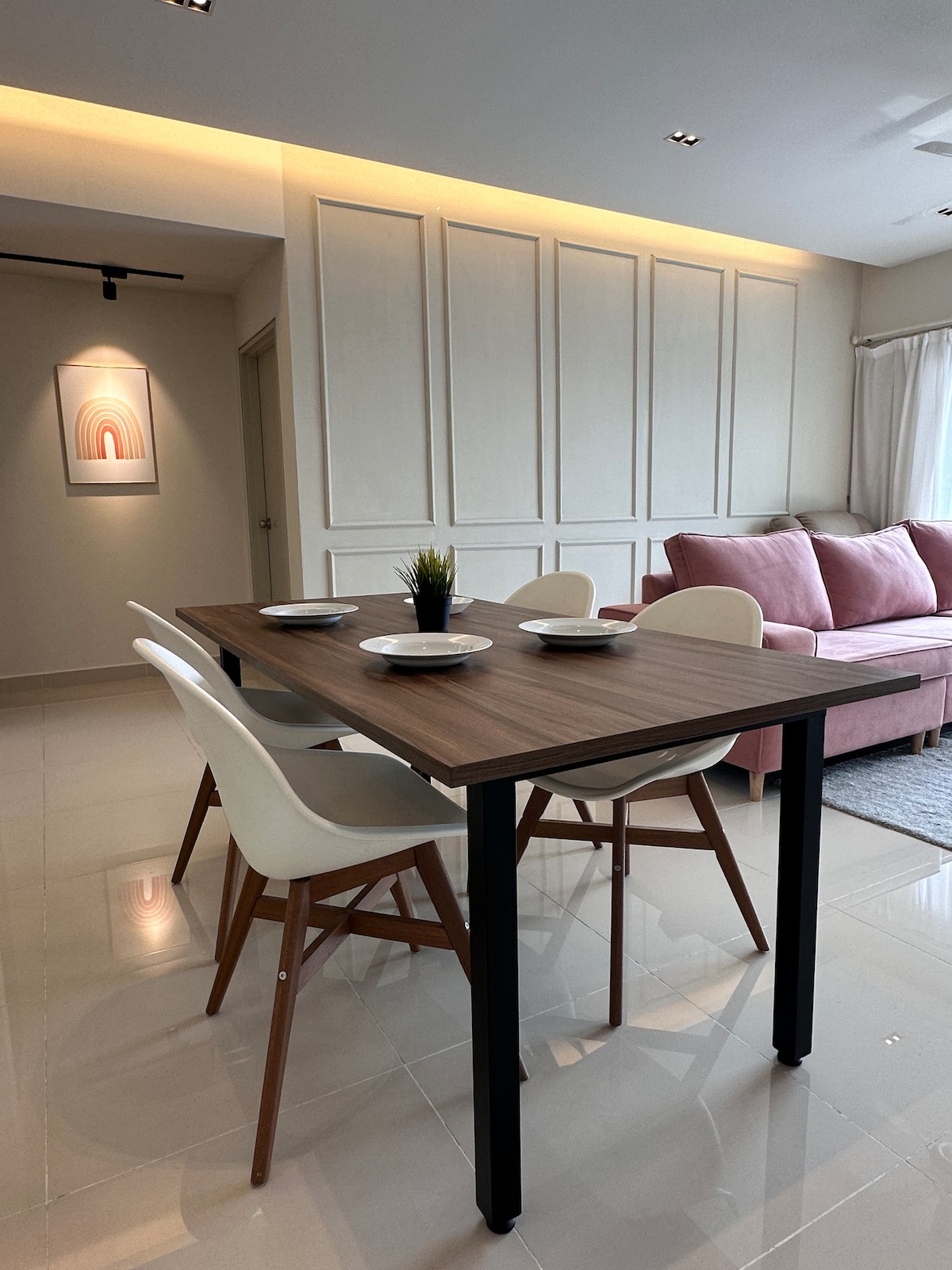 A dining area is presented with a wooden table surrounded by four white chairs, positioned against a backdrop of light-colored wall panels. A pink sofa is visible nearby, complemented by soft lighting and a piece of wall art above the table.