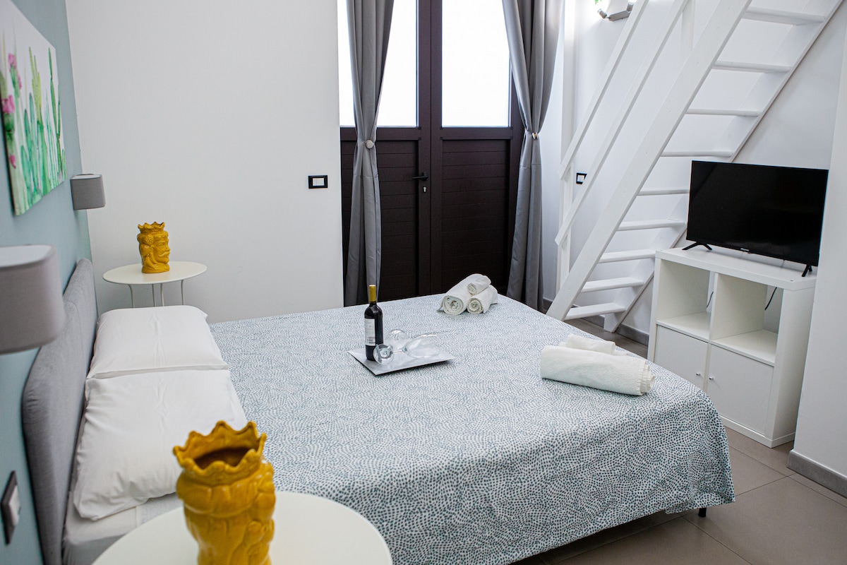 A spacious sleeping area features a large bed with a patterned cover and soft pillows. Natural light enters through tall windows draped with sheer curtains. A small table with a yellow vase adds a pop of color, while a sleek television is mounted nearby.