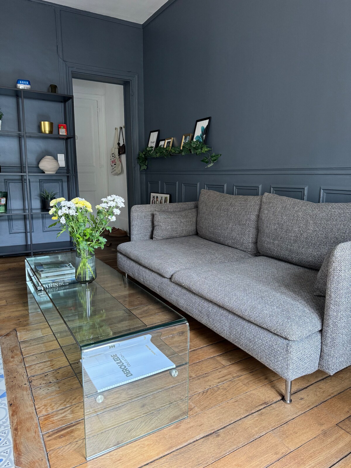 A modern living space showcases a gray fabric sofa paired with a clear glass coffee table. A bouquet of white flowers sits on the table. A shelving unit in the background displays decorative items and framed photos, against a dark blue accent wall.