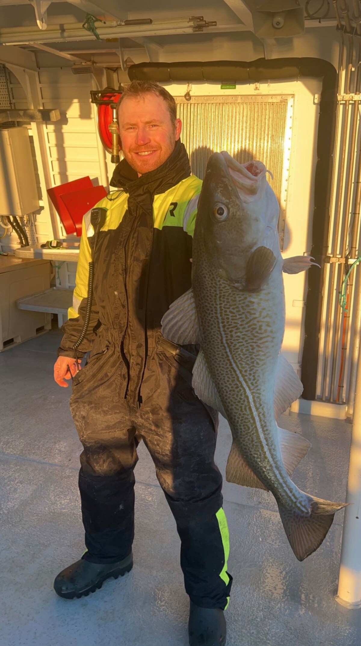 A person stands holding a sizable fish, showcasing its distinct coloration and features. The individual is dressed in dark fishing attire with bright accents, and the background includes a well-lit boat interior, emphasizing the experience of fishing trips.