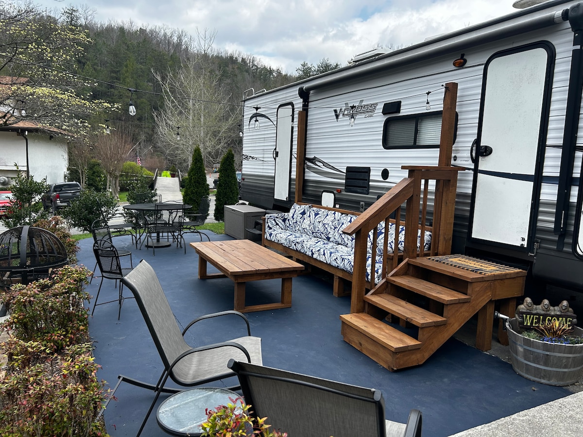 An outdoor seating area is set up next to the RV, featuring a wooden table and chairs, with a patterned sofa extending along the side. Steps lead up to the entry of the RV, and lush shrubbery surrounds the paved space, creating a cozy atmosphere.