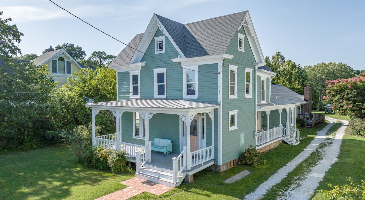 The exterior of a Victorian farmhouse is shown, featuring a light blue color with white trim. A welcoming front porch, supported by white columns, offers ample space for outdoor seating. Lush greenery surrounds the property, providing a serene environment. The driveway is visible on the right.