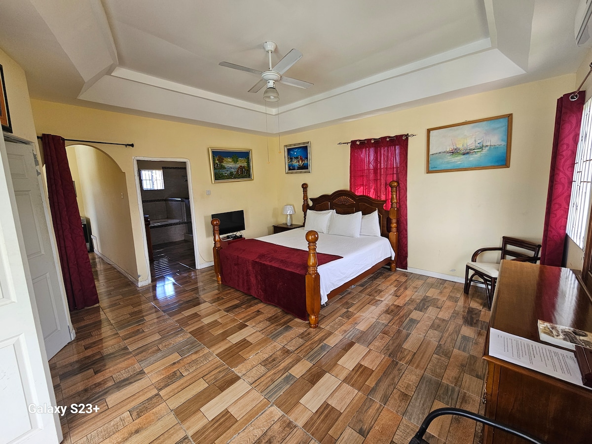 A spacious bedroom is furnished with a large wooden bed, accompanied by white linens and decorative pillows. A window with sheer burgundy curtains allows natural light to enter. Artwork adorns the walls, and a small desk and chair are positioned next to a flat-screen television.