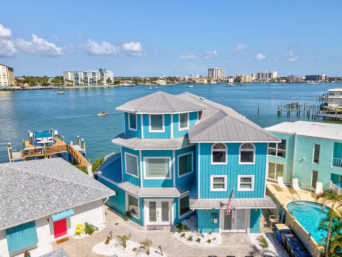 A large waterfront home is shown with a vibrant blue exterior, featuring multiple levels and distinctive architectural design. The calm water and distant shoreline are visible, highlighting the tranquil setting. Surrounding areas include a spacious patio and a functioning dock for relaxation.