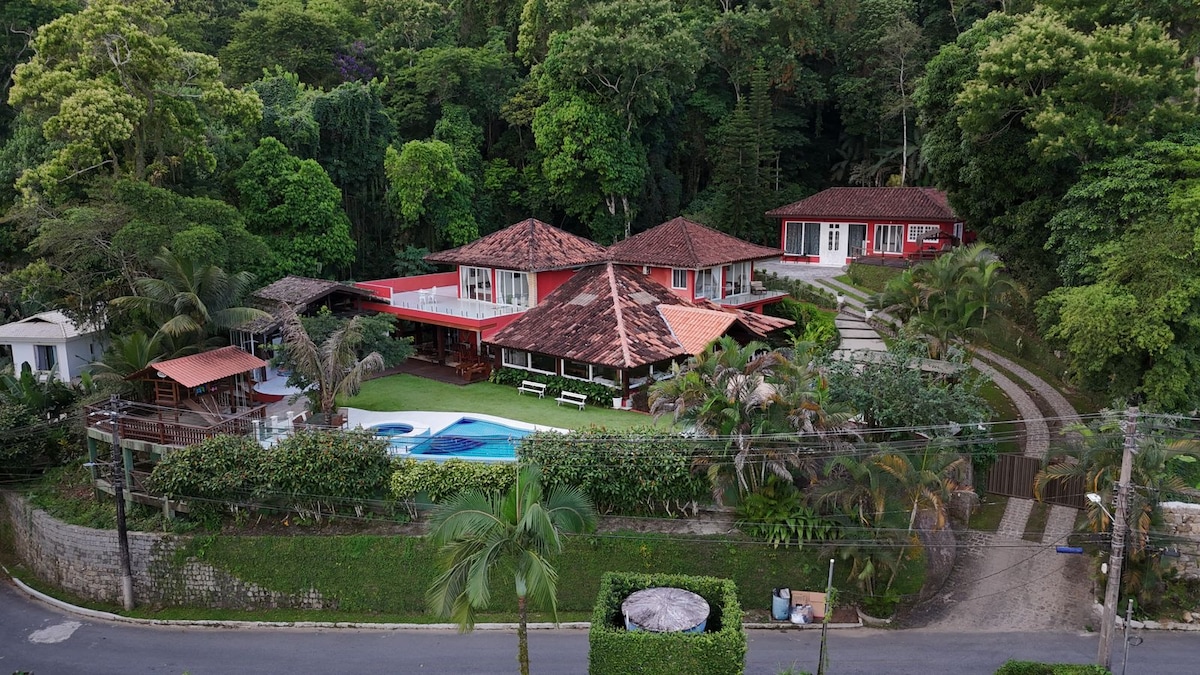 An aerial view showcases a spacious property nestled within a lush, green landscape. The house features distinctive red roofing, surrounded by gardens and outdoor seating areas. A pool is visible, along with well-maintained pathways leading to various sections of the home.