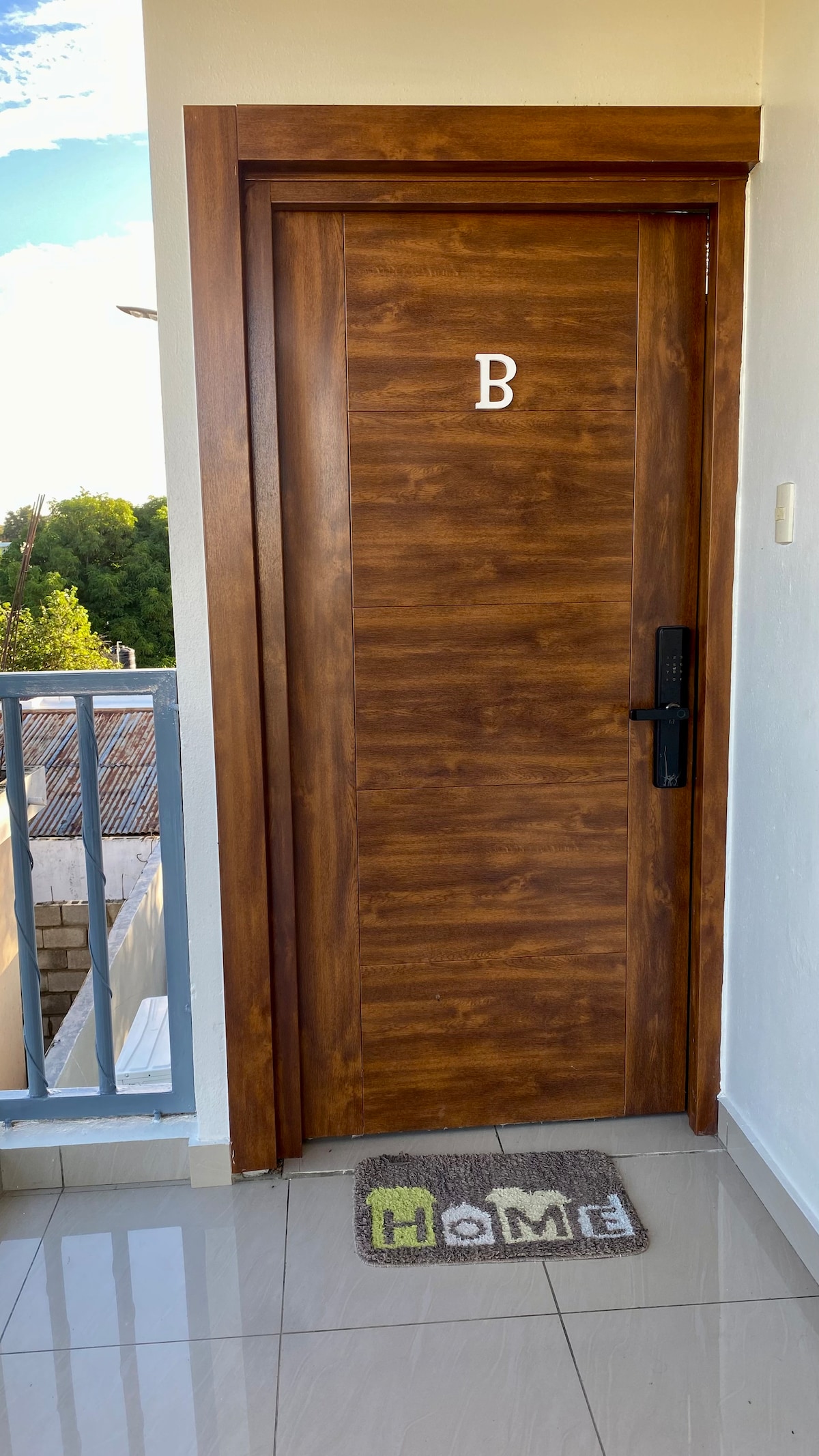 The image displays a wooden door marked with a letter 'B' at its center. A welcome mat with the word 'Home' is situated in front of the door on a glossy tiled floor. A glimpse of greenery is visible through the railing in the background.