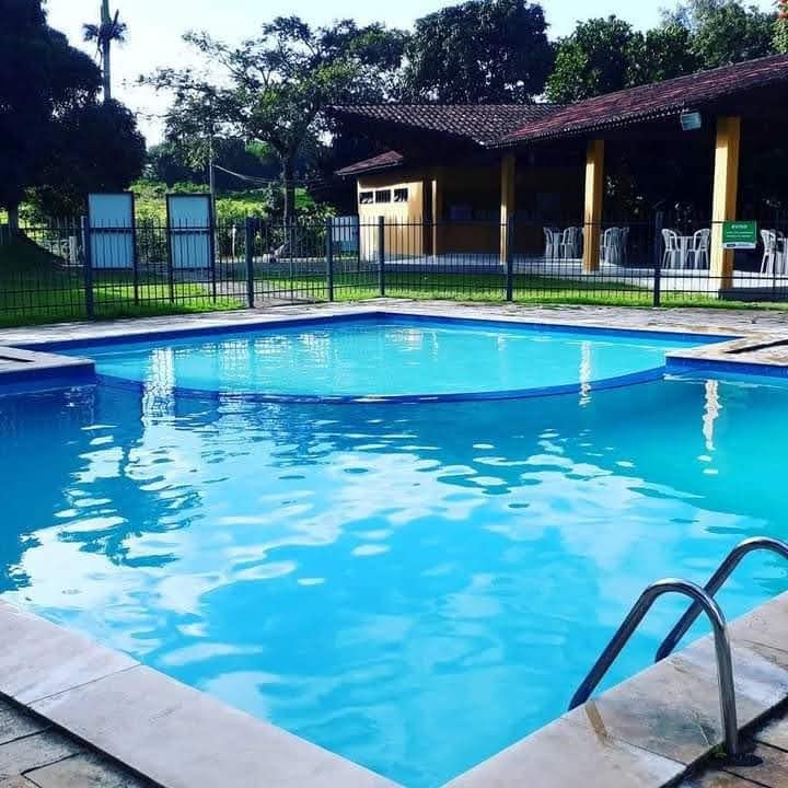 A swimming pool is surrounded by a secure fence, featuring clear blue water that reflects the sunlight. Lounge chairs are placed nearby on a tiled deck, with an open pavilion visible in the background, offering shaded seating for relaxation.