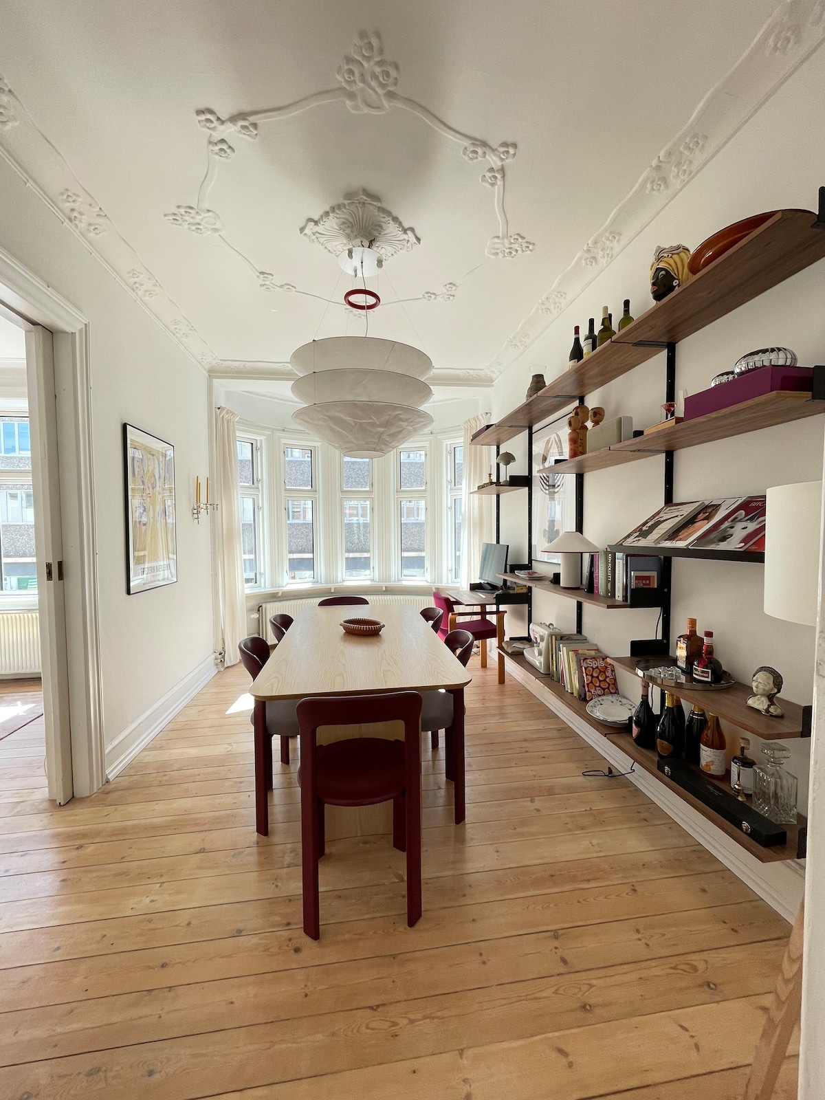 A dining area features a long wooden table surrounded by dark red chairs. A decorative shelf displays books and various items along one wall. Natural light floods the space through large windows, highlighting the ornate ceiling details and wooden flooring.