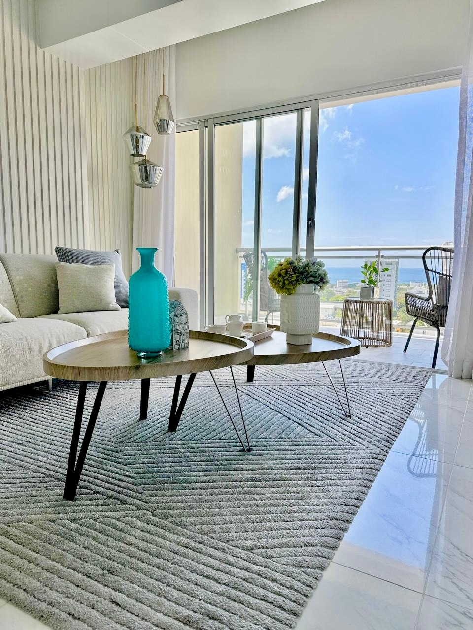A modern living area features a light-colored sofa positioned beside two small, round coffee tables. A turquoise glass vase adorns one table, while a potted plant is seen on the other. Floor-to-ceiling windows allow natural light, with a view of distant greenery and the sea.