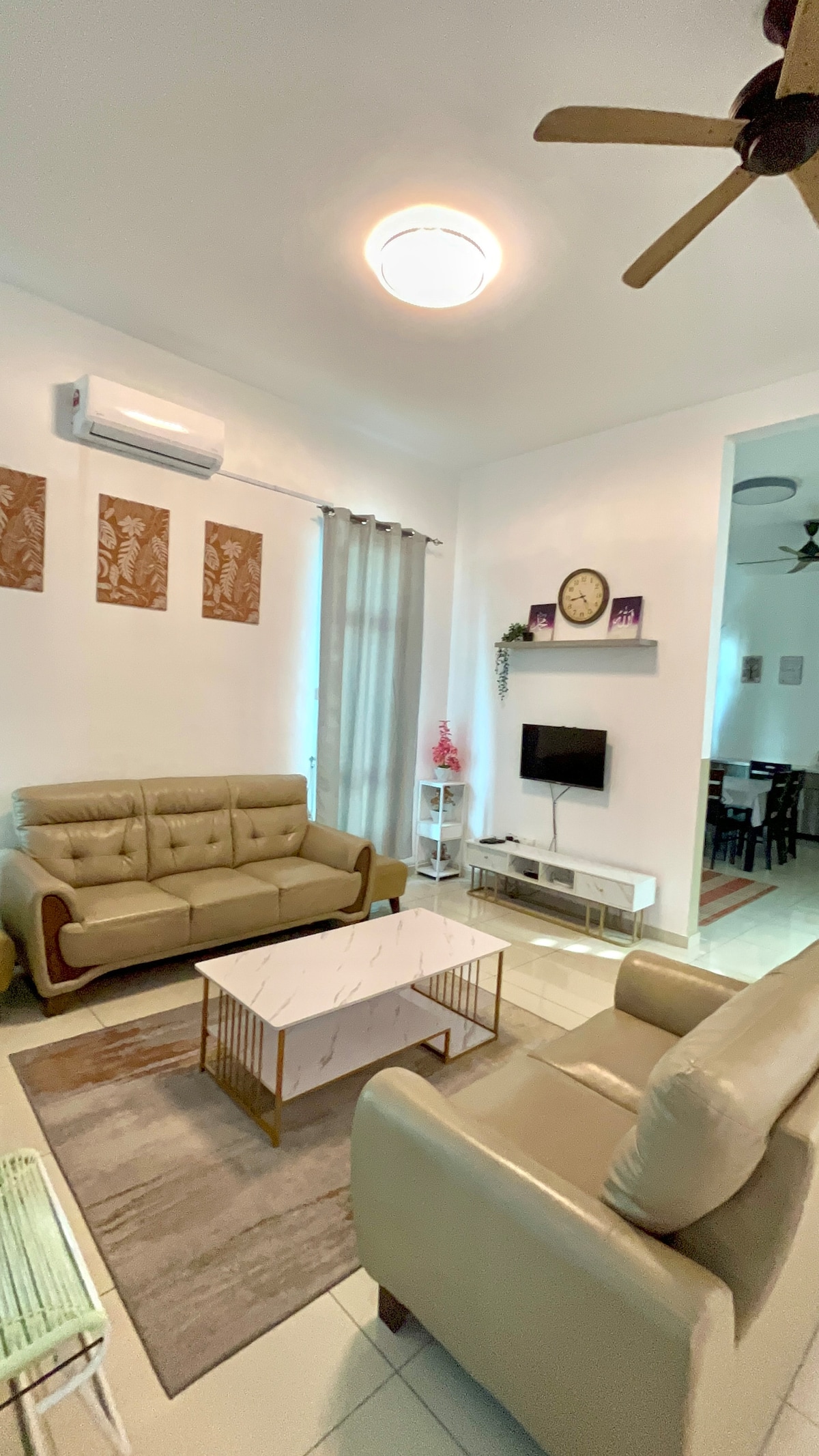 A well-lit living area features a minimalist design with beige sofas and a marble coffee table at the center. Decorative wall art is displayed above the seating, and a flat-screen television is mounted on the wall. Natural light filters in through sheer curtains.