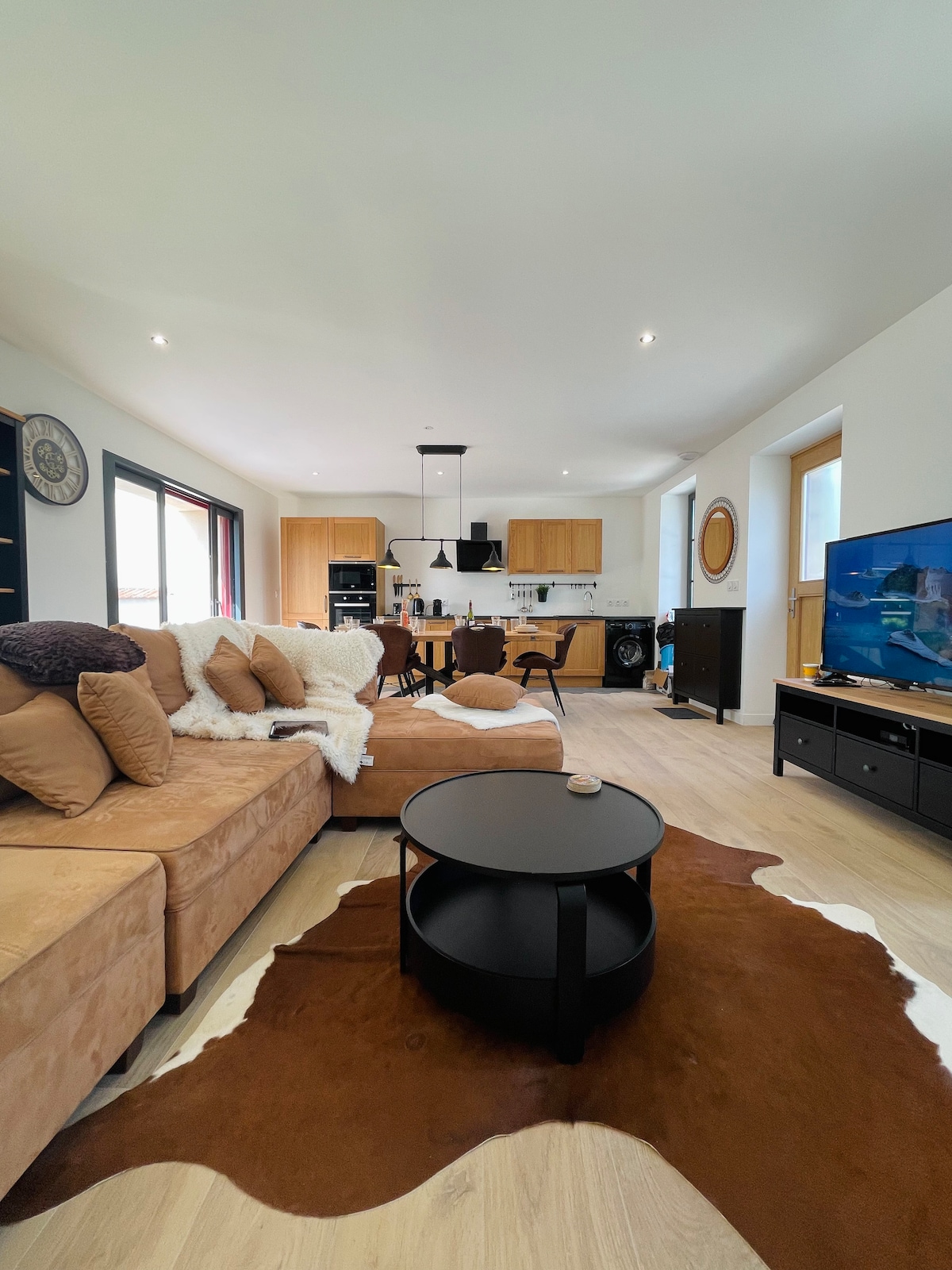 A spacious living area features a large, curved sofa adorned with decorative pillows and a throw blanket. A circular coffee table sits in front, complemented by a television on a stand. The open kitchen is visible in the background, enhancing the overall inviting space.
