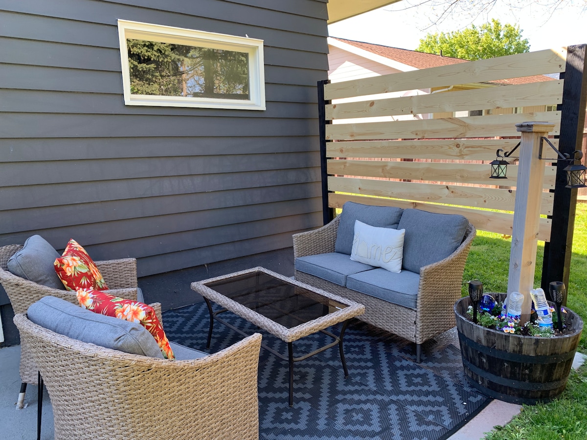 A cozy outdoor seating area is presented, featuring a gray sofa and two matching chairs. A glass-top coffee table sits in the center, complemented by decorative throw pillows. A wooden privacy screen is visible in the background, along with a garden space and a decorative planter.
