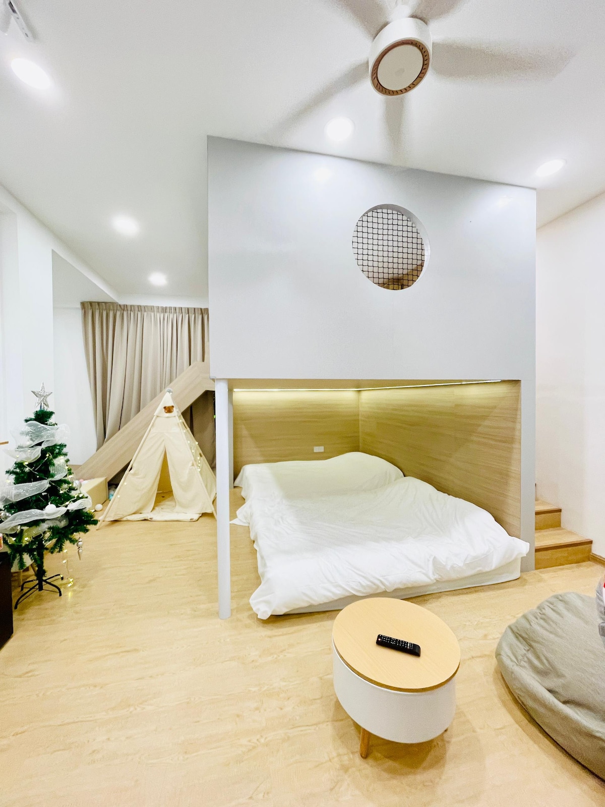 A cozy sleeping area features a spacious bed positioned against a wooden backdrop. A playful tent is located in the corner, adding a fun touch. Soft lighting creates a warm ambiance, while a small round table and a potted tree contribute to the inviting environment.