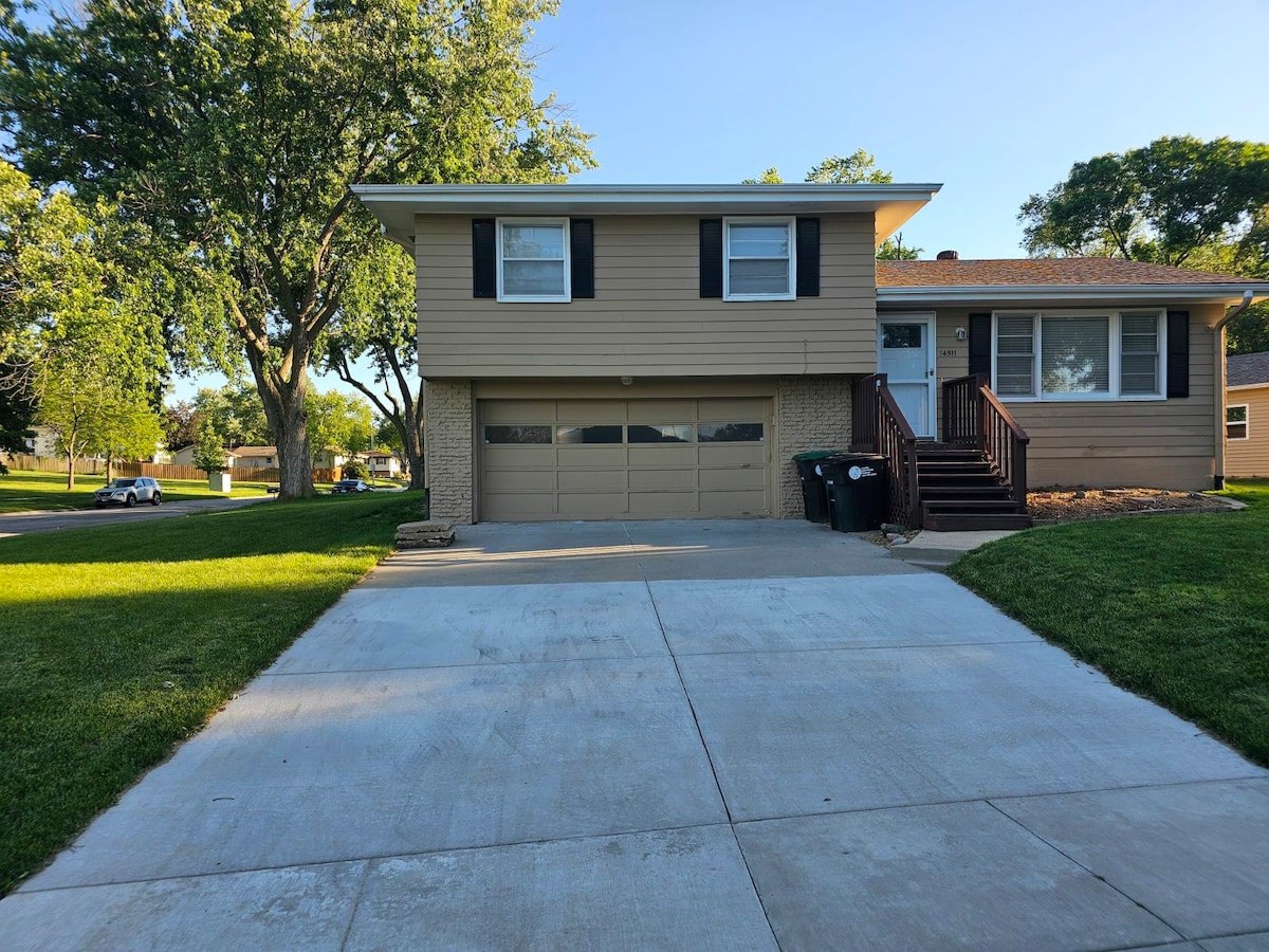 A two-story home is shown, featuring a beige exterior and a brown front door. The driveway leads to a two-car garage. Lush green grass surrounds the yard, with trees providing shade nearby. The sunlit street and peaceful neighborhood are visible in the background.
