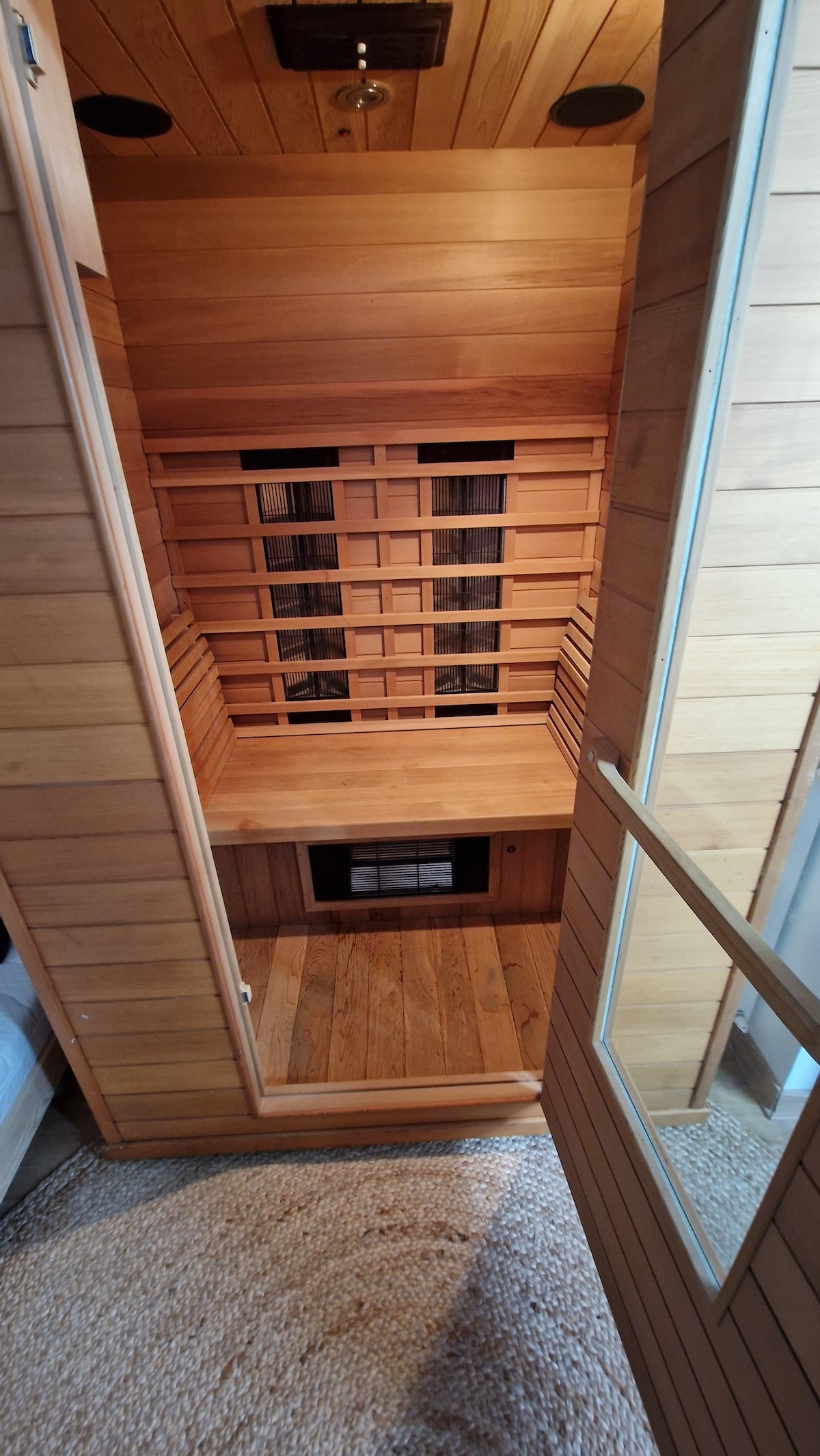A wooden infrared sauna features a spacious interior with slatted wooden seating. The walls are lined with warm wood paneling, and a large glass door allows for easy access. Natural light filters in through a small window, enhancing the soothing atmosphere.