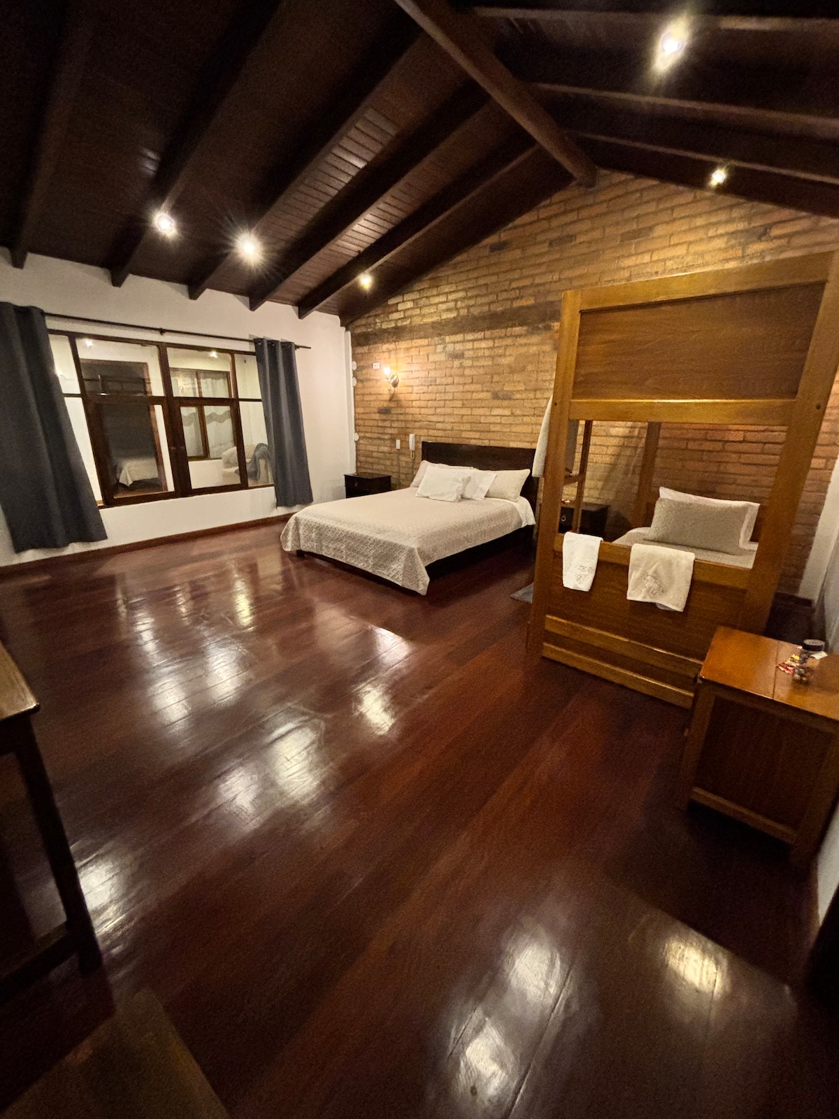 A spacious bedroom features wooden beams overhead, complemented by a warm brick wall. The room includes a large bed with white bedding and is illuminated by soft overhead lighting. A window with curtains allows natural light, and wooden furnishings are arranged thoughtfully.
