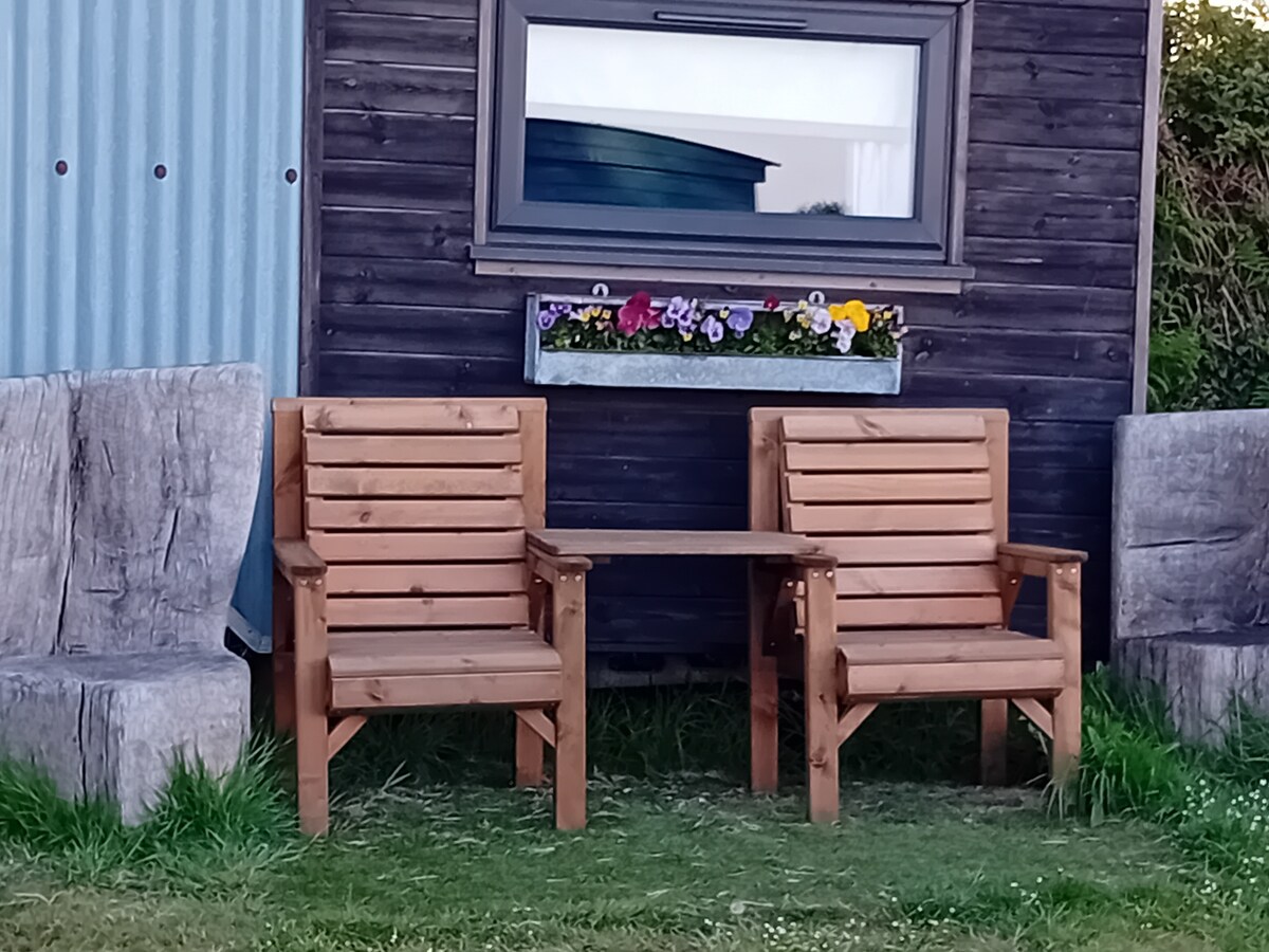 Two wooden armchairs and a small table are positioned beside the hut. A planter filled with colorful flowers is mounted under the window, enhancing the outdoor seating area, which is framed by grassy terrain.