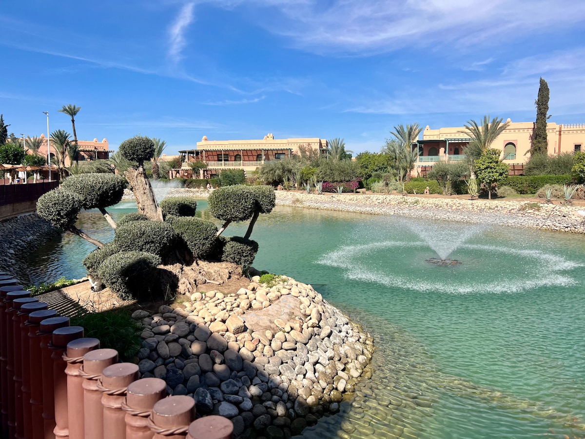 A serene landscape features a calm body of water with a central fountain creating gentle ripples. Lush greenery, including sculpted trees, surrounds the water, while well-maintained pathways and distant palm trees enhance the tranquil environment.