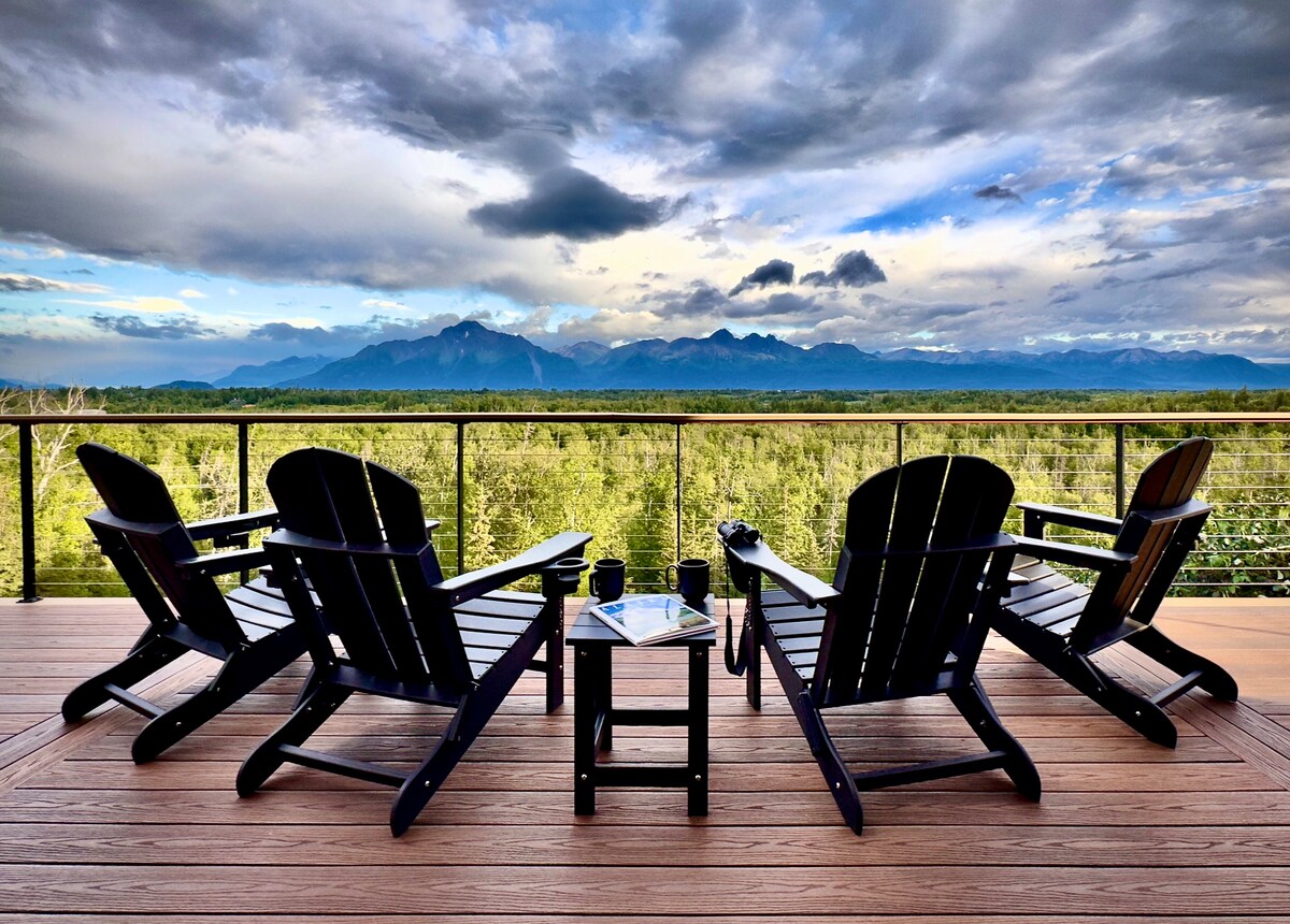Four Adirondack chairs are arranged around a small table on a spacious wooden deck. A panoramic view of lush greenery and distant mountains is visible under a dramatic sky filled with clouds, offering a serene outdoor setting.