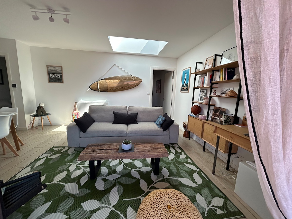 A light-filled living area is showcased with a spacious sofa adorned with dark cushions. A wooden coffee table sits on a patterned rug, while a surfboard is displayed on the wall above. A large skylight provides ample natural light, enhancing the room's modern decor.