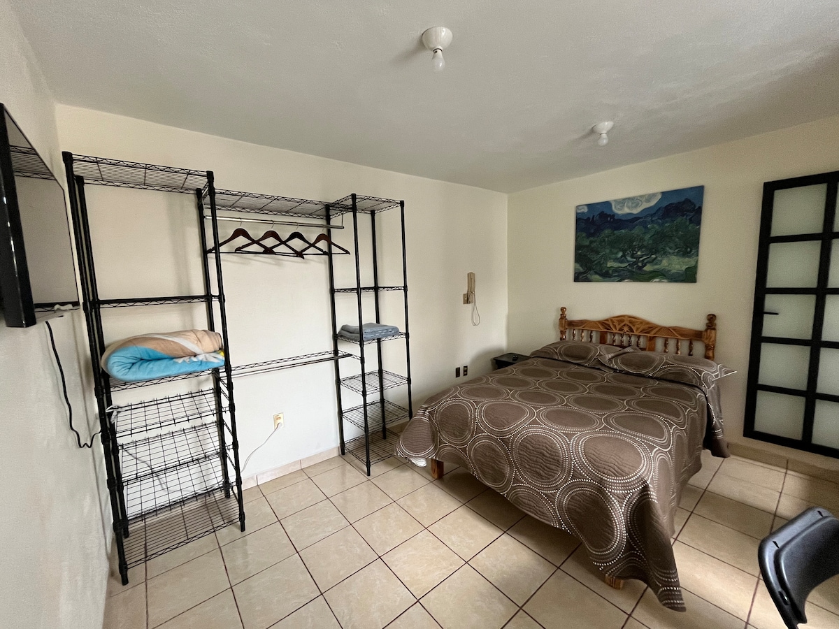 A spacious guest room is presented, featuring a matrimonial bed with a neutral-colored blanket. A shelving unit with hangers and towels is visible, alongside a flat-screen TV mounted on the wall. Natural light enters through a window, illuminating the room's simplicity.