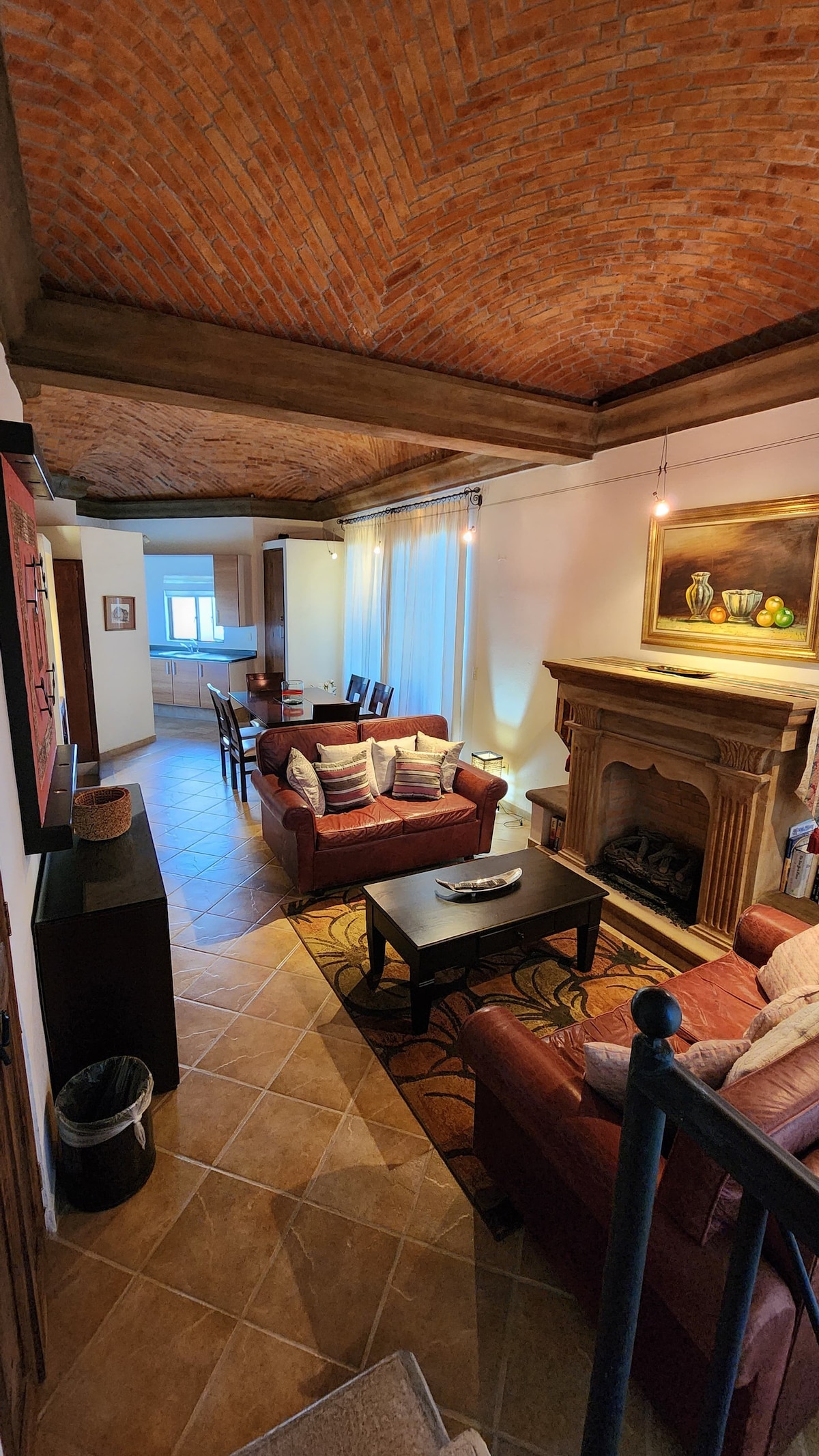 A spacious living area features red sofas and a coffee table centered around a stone fireplace. A dining area with a table and chairs is visible in the background, along with kitchen space. Soft lighting highlights the vaulted brick ceiling, enhancing the cozy ambiance.