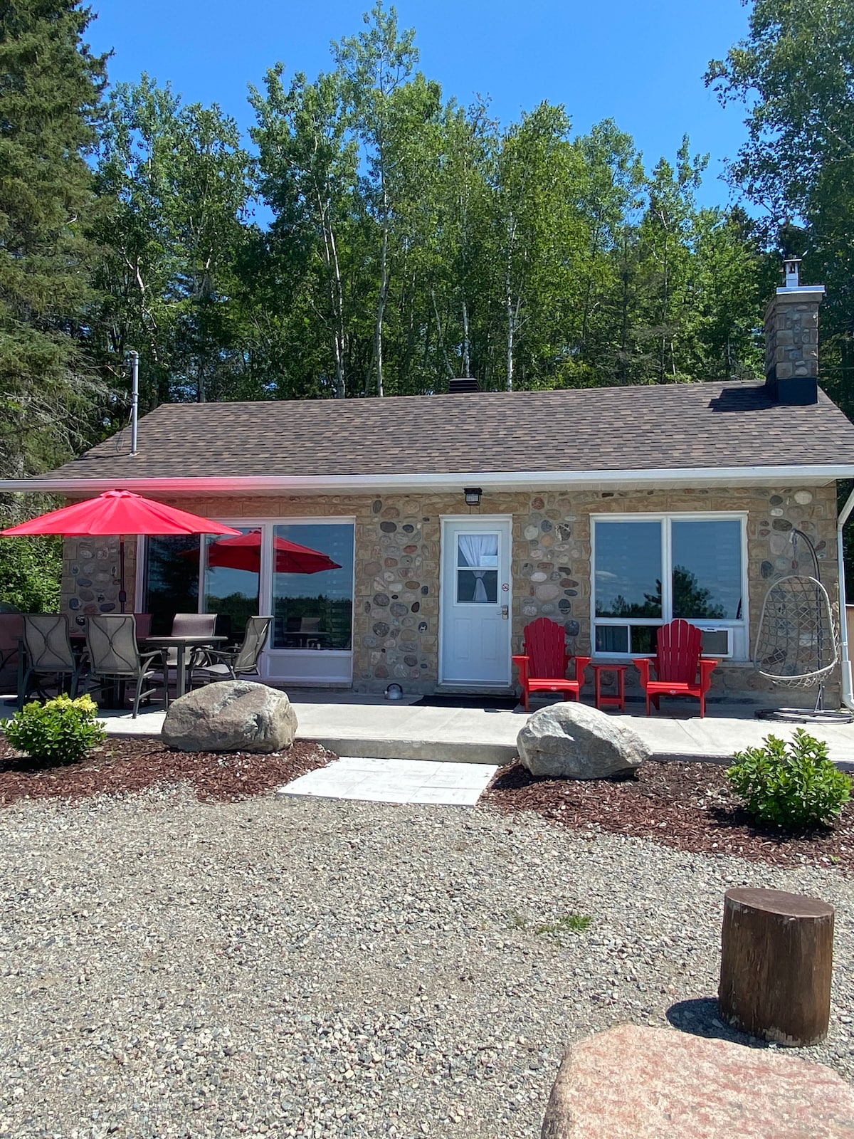 A charming stone cottage is set against a backdrop of lush trees. A patio area features red umbrellas over outdoor seating, with two red chairs positioned on a stone walkway. Surrounding green foliage and rocks enhance the cottage's natural setting.