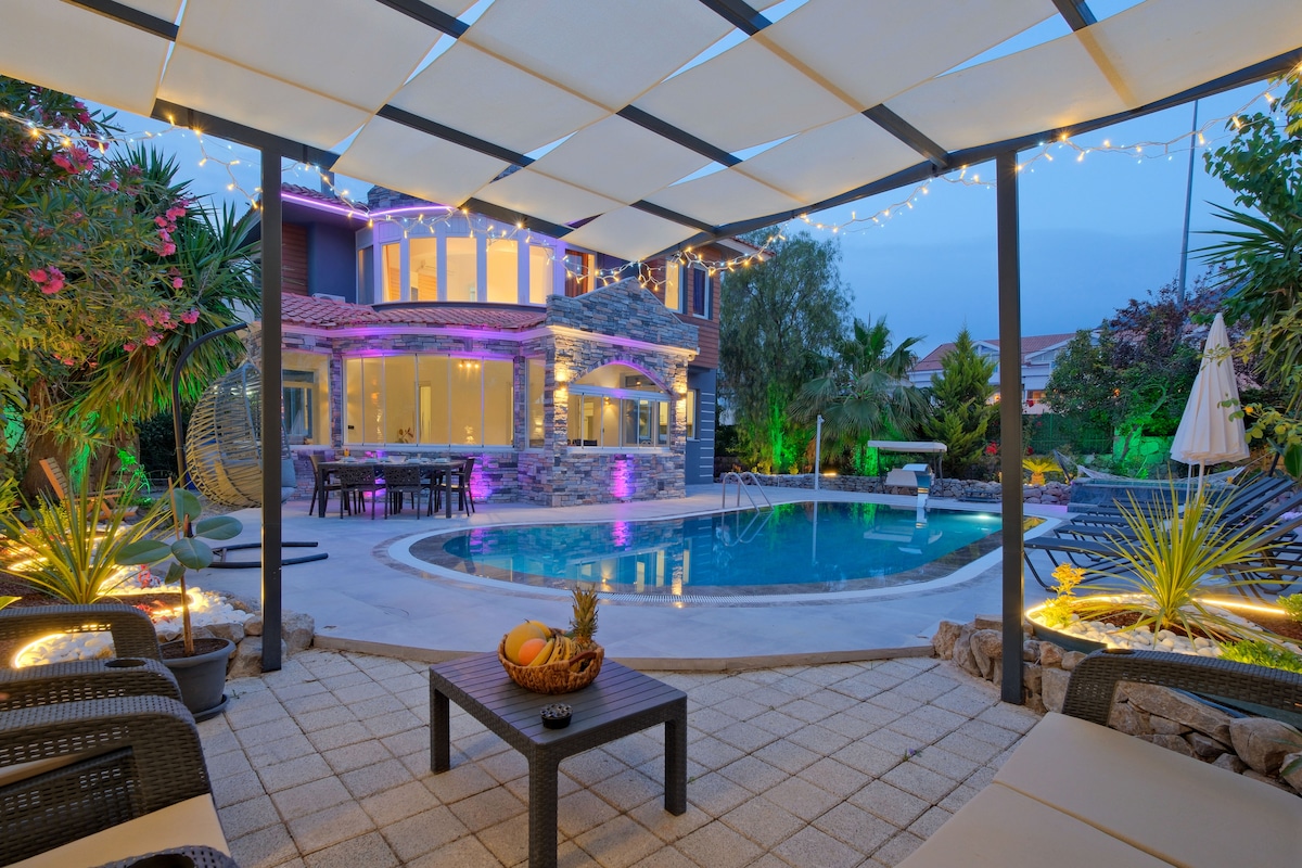 An outdoor area features a private pool surrounded by a spacious patio with lounge furniture. Soft lighting illuminates the surroundings, accentuating lush greenery and decorative plants. A table with a fruit bowl sits in the foreground, creating a welcoming space for relaxation and entertainment.