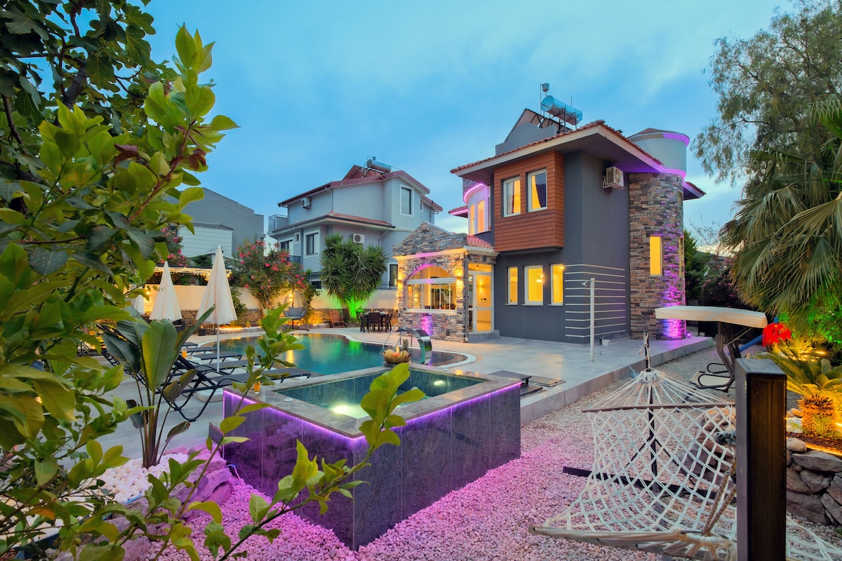 A modern villa is surrounded by lush greenery, showcasing a spacious private pool illuminated with soft lighting. A hammock is positioned nearby on a bed of pebbles. The two-story structure features large windows and an inviting patio area, enhancing the outdoor living experience.