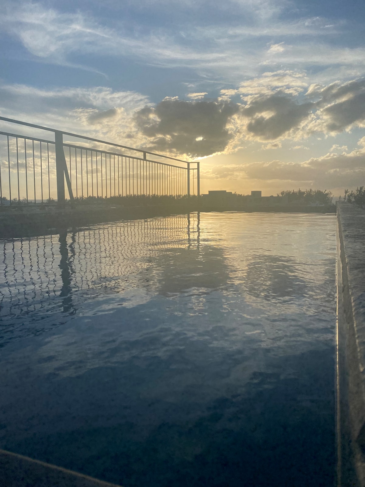 The rooftop swimming pool reflects the warm hues of the sunset, surrounded by a railing that provides safety and scenic views. The calm water mirrors the sky, creating a serene atmosphere perfect for relaxation.