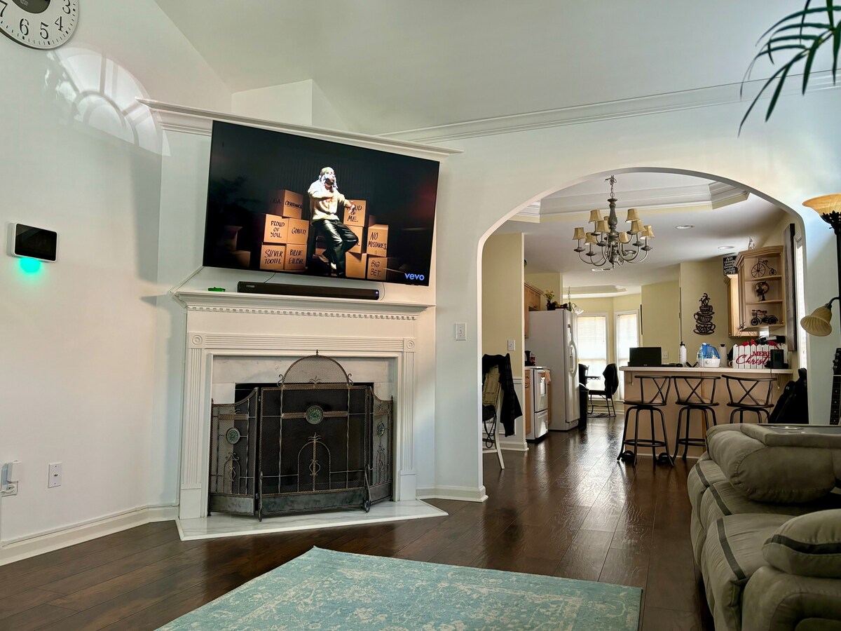 A spacious living area features a mounted television above a classic fireplace, surrounded by decorative boxes. Natural light enters through an archway leading to a modern kitchen with a dining area. Comfortable seating is provided by a light-colored sofa, enhancing the inviting space.