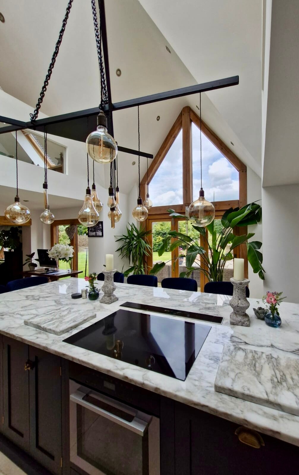 A modern kitchen features a marble island with an integrated cooktop. Pendant lights with glass globes hang above, illuminating the space. Large windows allow natural light to fill the area, showcasing indoor plants and a dining table set against a backdrop of greenery.