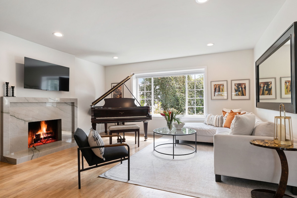 A light-filled living room features a grand piano beside a sleek marble fireplace. A comfortable sectional sofa and a modern glass coffee table invite relaxation, while framed artwork adorns the walls. Large windows provide views of the landscaped outdoor space.