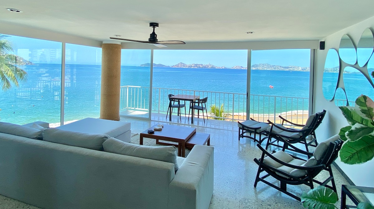 A light-filled living area features large glass windows offering unobstructed views of the ocean. Comfortable seating arrangements include a spacious sofa and lounge chairs. A wooden coffee table sits in the center, complemented by a ceiling fan that enhances airflow.