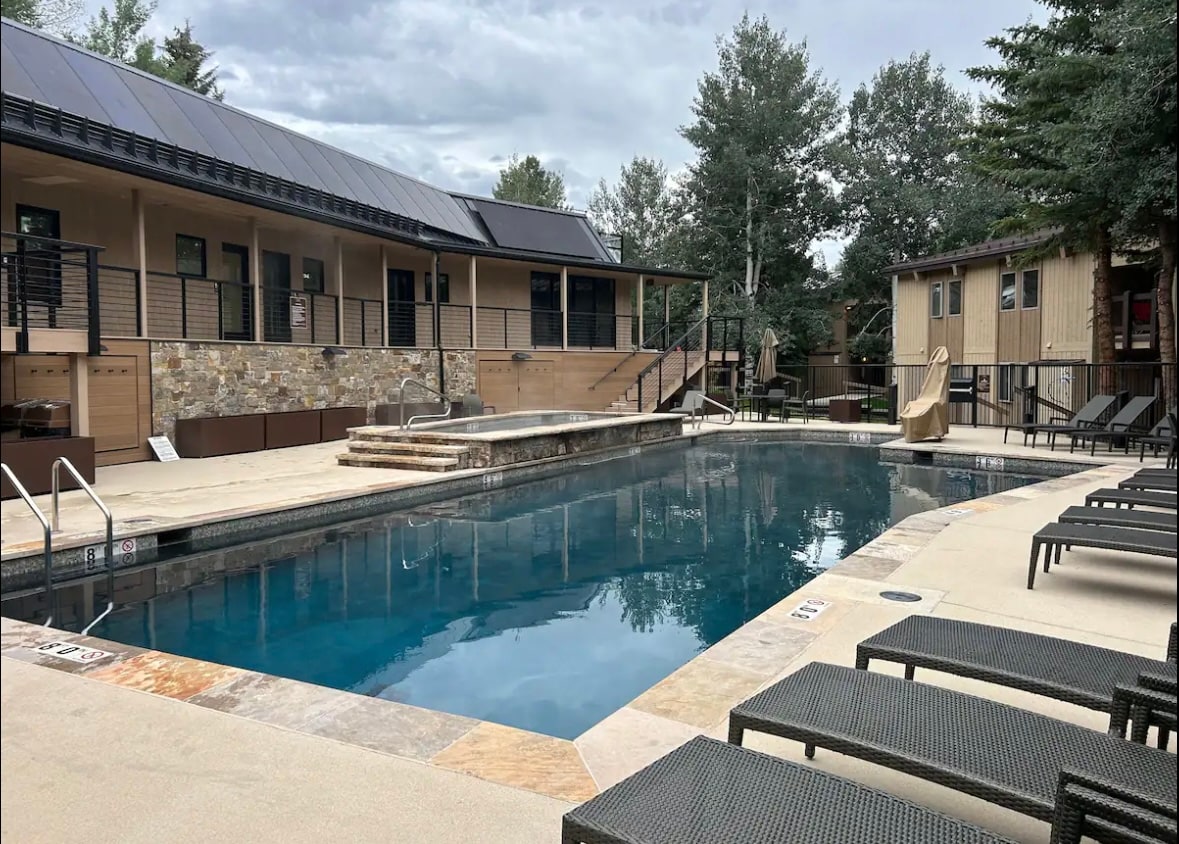 An outdoor pool area is highlighted, featuring a clear, blue swimming pool surrounded by lounge chairs. Steps lead up to a stone patio, while trees provide a natural backdrop. The building is visible in the background, with multiple levels and outdoor lighting.