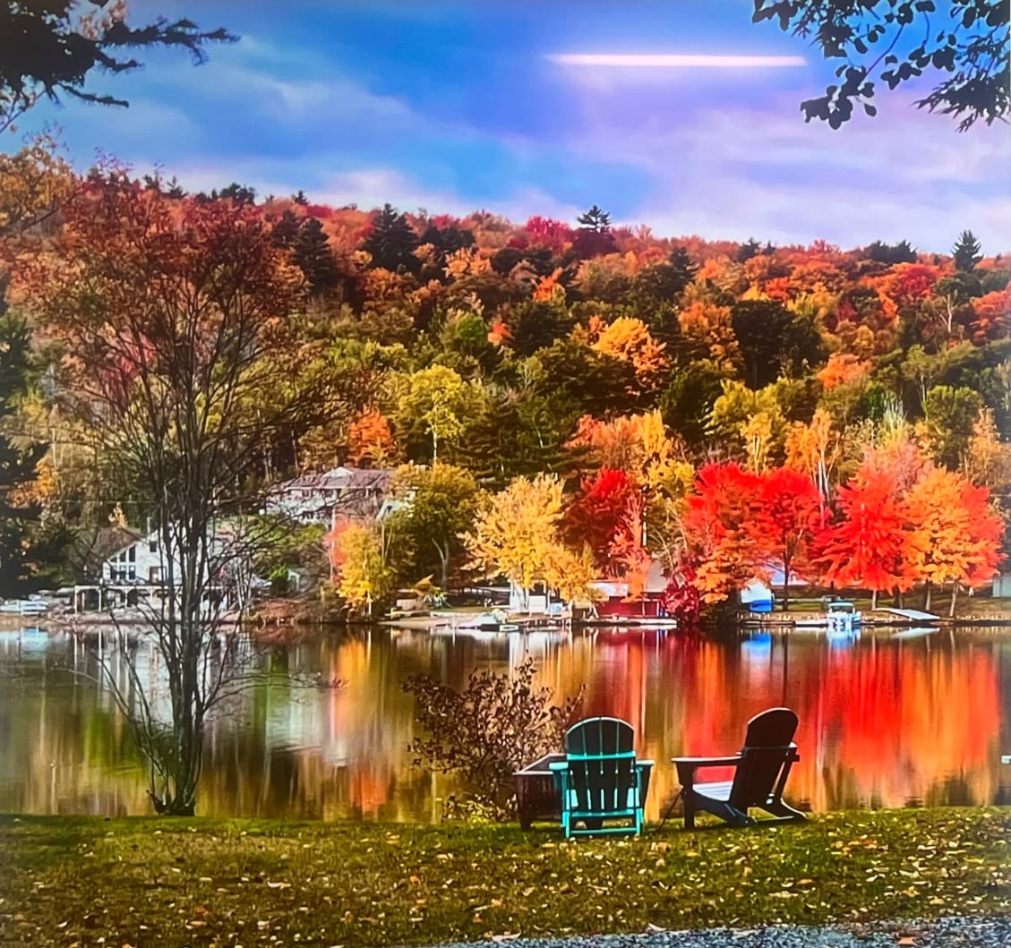 A serene lakeside view is presented, featuring vibrant autumn foliage reflected in the calm water. Two adirondack chairs are positioned on a grassy area, inviting relaxation. A private dock and several boats are visible along the shore, framed by a backdrop of colorful trees.