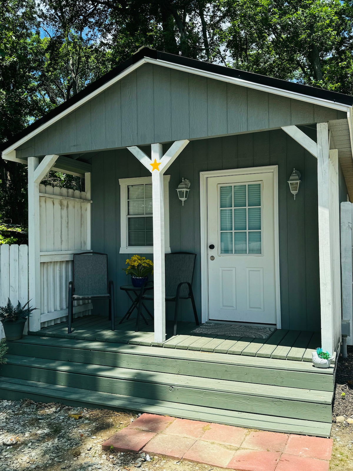 A charming tiny home entrance is presented, featuring a light grey exterior with decorative white trim. Two chairs with cushions sit beside a small potted yellow flower. A front door with a window is centered, and the porch is framed by a white railing.