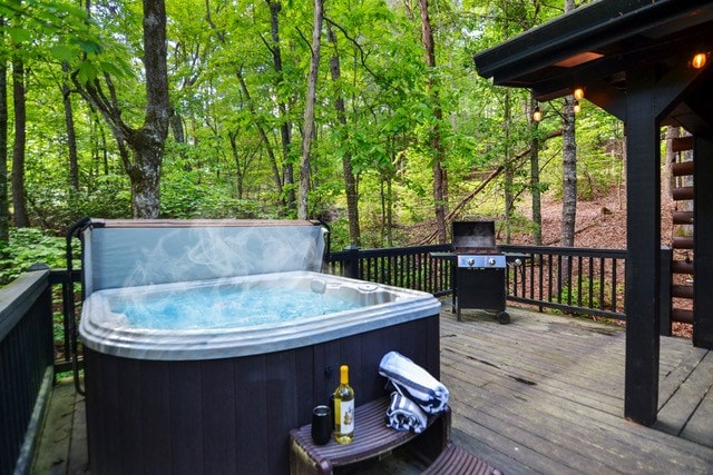 A private hot tub is positioned on a wooden deck surrounded by lush foliage. A grill can be seen nearby, and a bottle of wine sits with two glasses on a small table. The serene setting and gentle steam rising from the hot tub create a relaxing ambiance.