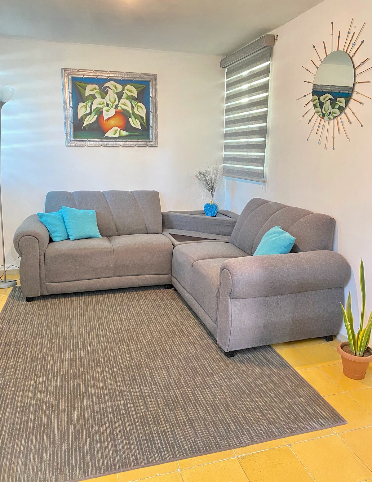 A gray sectional sofa is positioned in a bright living area. Two turquoise accent pillows are placed on the sofa, complementing a large wall art piece. A round mirror hangs above the seating, while a small potted plant adds a touch of greenery to the space.