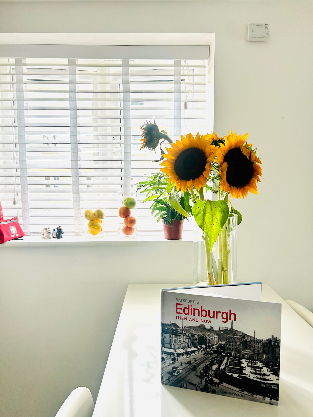 A modern dining space is highlighted by a vase of sunflowers, adding a touch of brightness. A book about Edinburgh is placed on the table, with natural light filtering through the slatted blinds. Decorative fruit and a small model are visible on the windowsill.