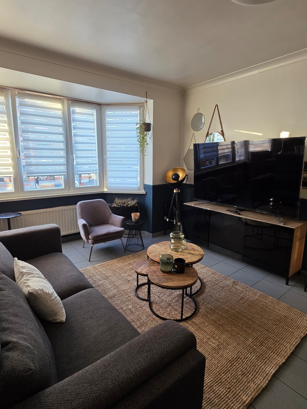 A cozy living area features a dark sofa, paired with a light purple chair. Two round coffee tables are positioned in front, accompanied by a woven rug. A large television is mounted on the wall, while sunlight filters through the window adorned with shutters.