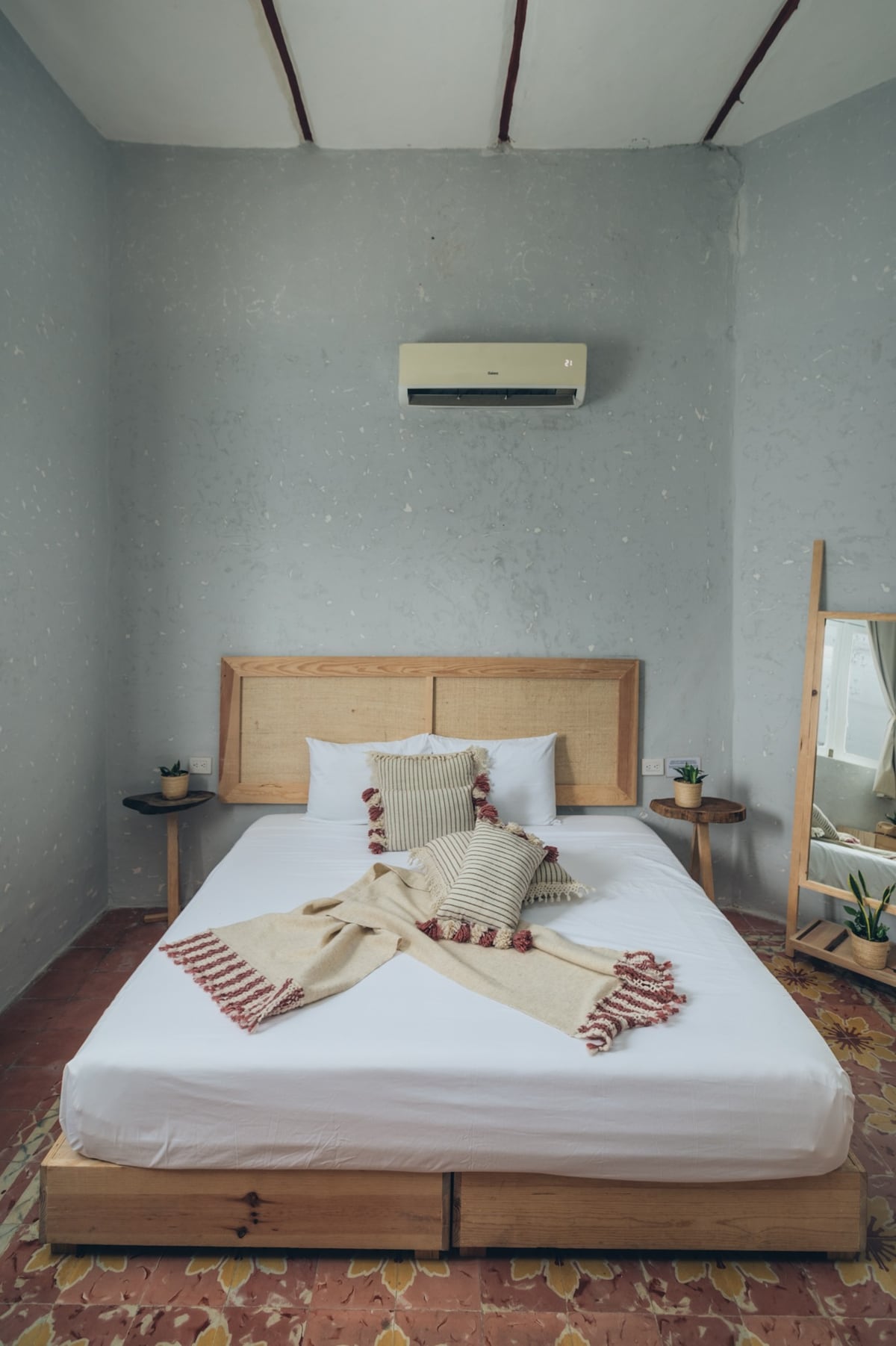 A cozy bedroom features a spacious bed adorned with decorative pillows and a woven throw blanket. A wooden headboard complements the textured, light gray walls. Small potted plants are positioned on either side, and a full-length mirror reflects the natural light coming from the nearby window.