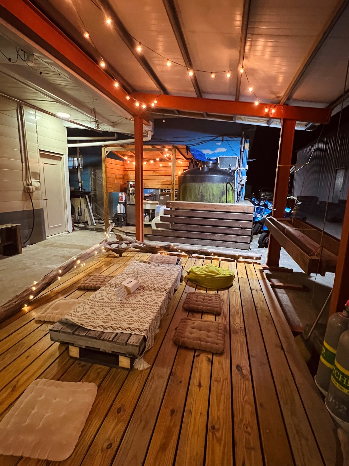 An outdoor space features a wooden deck illuminated by warm string lights. Mats and cushions are arranged on the floor, with a wooden bench positioned nearby. A backdrop of simple structures is visible, contributing to a cozy atmosphere.